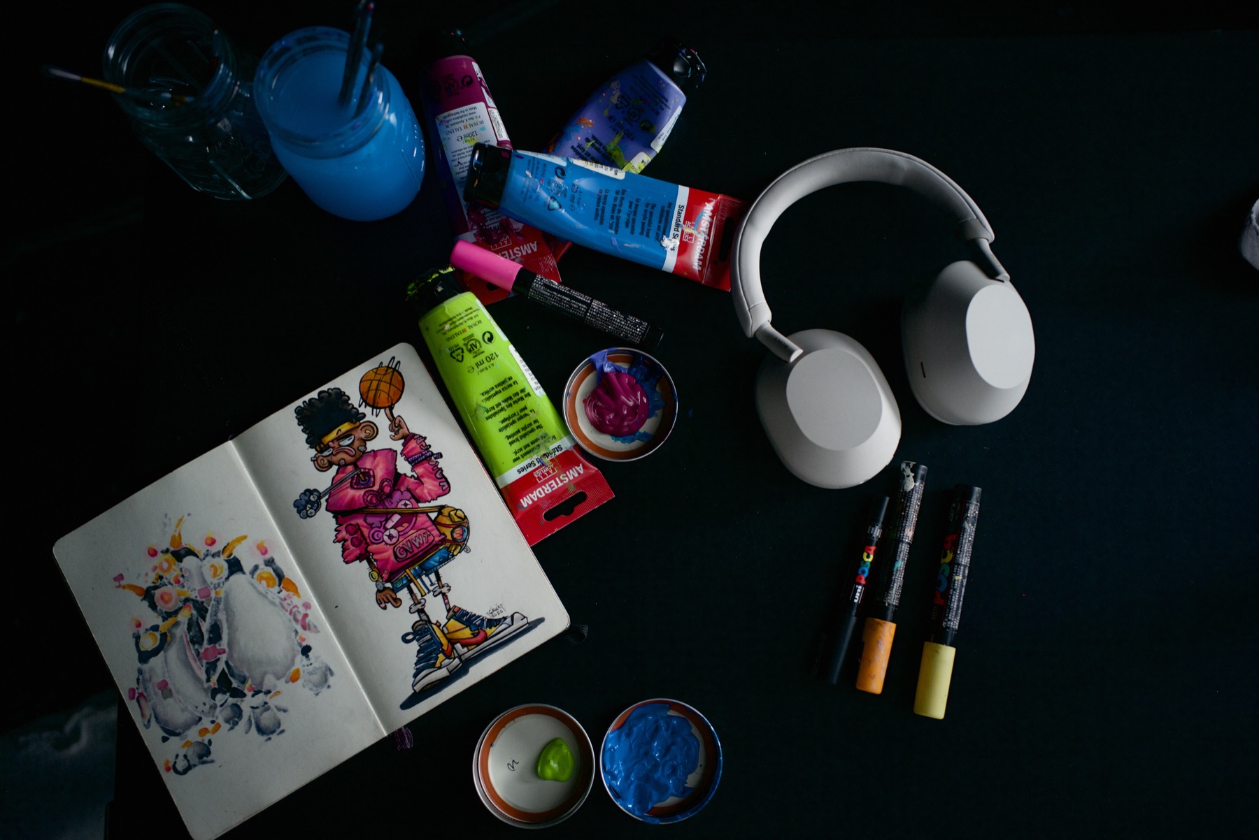The tools, flat lay: sketchbook, paints, markers, and the WH-1000XM5